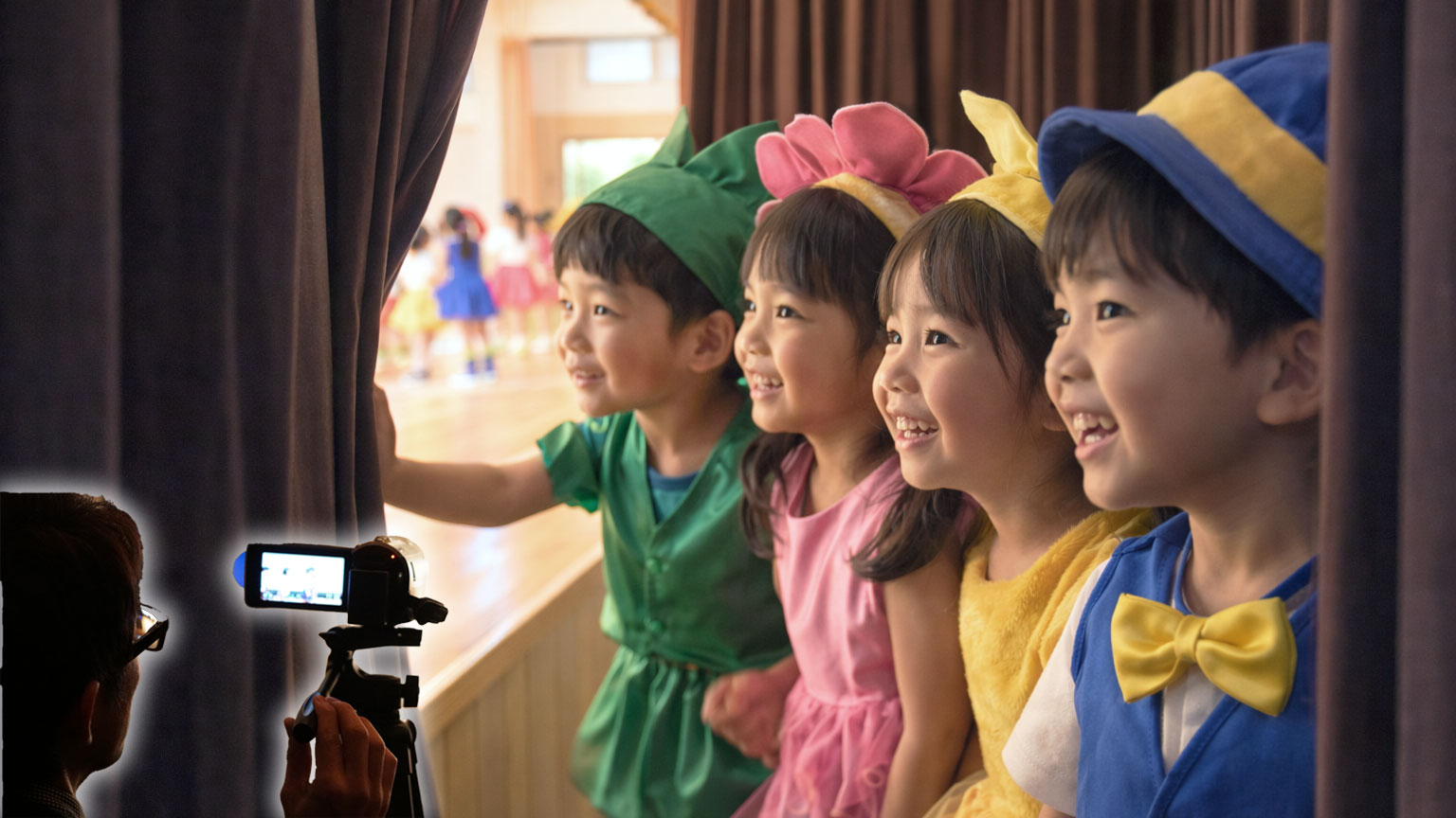 幼稚園の発表会で舞台袖からカラフルな衣装姿の園児たちをビデオカメラで撮影する撮影業者。カーテンの隙間から本番のステージを笑顔で見つめる子どもたち